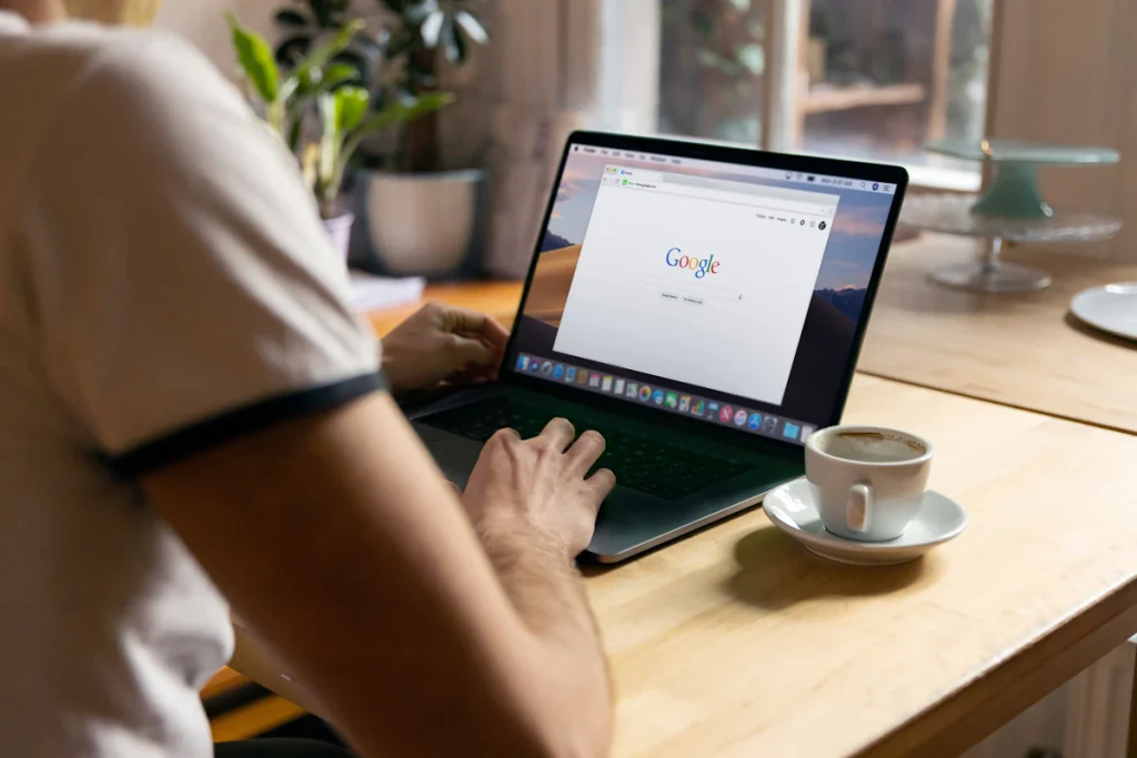 Man at a cafe table typing a query into the Google search page on a laptop
